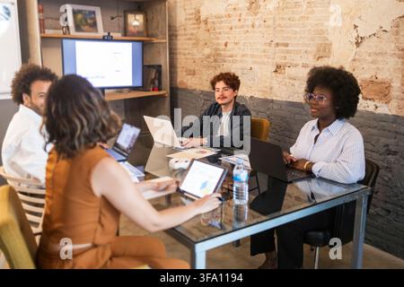 Des collègues divers utilisent des ordinateurs portables et des tablettes lors d'une réunion collaborative dans un bureau de coworking moderne avec des murs en briques apparentes et un espace de travail partagé. Banque D'Images