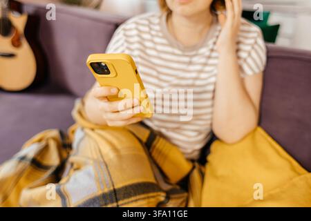 Jeune femme en t-shirt rayé assise sur un canapé à la maison, tenant un smartphone jaune tout en touchant ses cheveux. Une guitare et une couverture à carreaux créent un i Banque D'Images