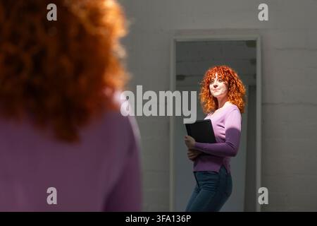 Jeune femme d'affaires rousse tenant une tablette dans un intérieur minimaliste, encadrée par une figure floue au premier plan et une forte lumière naturelle du soleil Banque D'Images