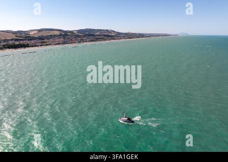 Vue aérienne d'un voilier naviguant sur la mer Adriatique turquoise près de la côte sablonneuse et du paysage urbain de Porto San Giorgio, Marches, Italie. Banque D'Images