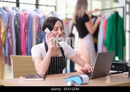 Jeune entrepreneur de mode parlant au smartphone tout en travaillant sur un ordinateur portable dans un atelier de vêtements avec des portants à vêtements, lumière naturelle vive, planification concentrée Banque D'Images