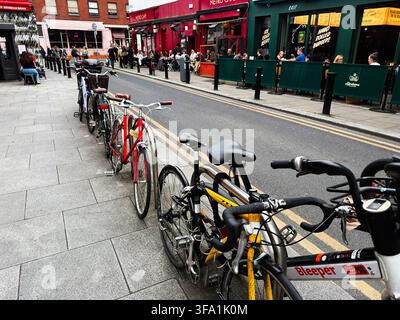 Dublin, Irlande – 22 mai 2026 : Des vélos garés bordent une rue animée de Dublin avec des cafés en terrasse, des restaurants et une atmosphère urbaine. Banque D'Images