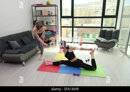 Deux femmes pratiquant des étirements et des poses de yoga sur des tapis d'exercice colorés dans un salon lumineux, une personne guidant une autre lors d'un entraînement à domicile à côté Banque D'Images