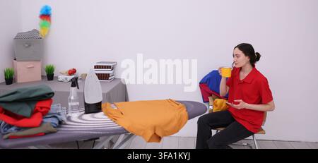 Jeune femme en chemise rouge assise à côté d'une planche à repasser avec du linge plié, tenant une tasse jaune et un smartphone dans une buanderie domestique lumineuse avec des vêtements Banque D'Images