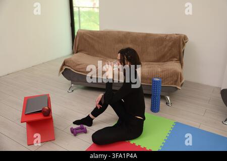 Femme en vêtements de sport noirs assise sur un tapis d'exercice coloré à la maison, buvant de l'eau à côté de haltères, un ordinateur portable, une pomme, un rouleau en mousse et un canapé dans une pièce lumineuse Banque D'Images