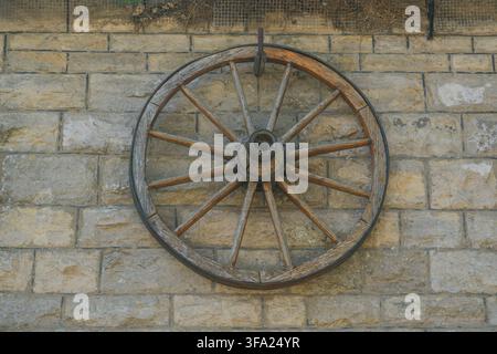 Close-up d'une roue de char en bois antique suspendue comme décoration sur un mur de briques en pierre jaune texturé sous la lumière naturelle du jour. Banque D'Images