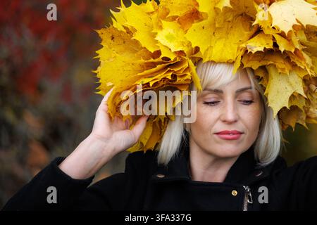 Un portrait en gros plan d'une femme blonde pensive tenant une couronne massive de feuilles d'automne jaune vif sur sa tête. Elle porte une veste noire contre un blü Banque D'Images