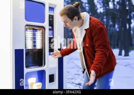 Jeune homme surpris regardant une machine à café dans un parc enneigé en hiver Banque D'Images
