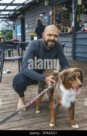 Un homme portant une combinaison de plongée agenouillé sur une terrasse en bois caresse son chien, un berger australien. Derrière eux, des gens se détendent dans un café au bord de la plage par une journée ensoleillée Banque D'Images