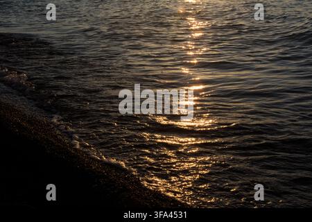 La lumière dorée du soleil se reflète sur les vagues douces d'une mer calme au coucher du soleil, projetant une lueur chaude sur le rivage de galets dans une atmosphère paisible et sereine Banque D'Images