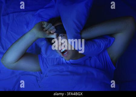 Un homme en colère portant un masque pour dormir couvre ses oreilles avec un oreiller et souffre d'insomnie, de bruit et de privation de sommeil dans une chambre bleu foncé la nuit Banque D'Images