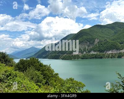 Un lac turquoise serpente à travers une vallée montagneuse, entouré de pentes vertes luxuriantes et encadré par la végétation sous un ciel nuageux. Banque D'Images