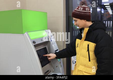 Jeune homme en veste d'hiver jaune vif et casquette utilisant un écran tactile, un distributeur de billets et retirant ou déposant de l'argent Banque D'Images