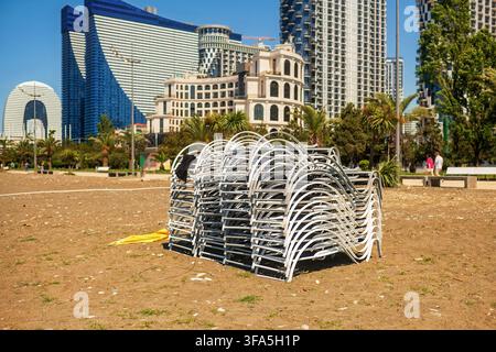 Transats en plastique empilés sur le rivage d’une station balnéaire de sable de galets avec en fond des gratte-ciel de ville modernes, des palmiers, une promenade sous un ciel ensoleillé. Banque D'Images