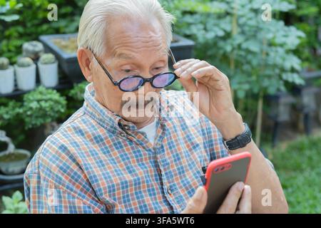 Homme asiatique âgé utilisant un smartphone, portant des lunettes et ayant une mauvaise vue en extérieur. Concept de problèmes de vue. Banque D'Images
