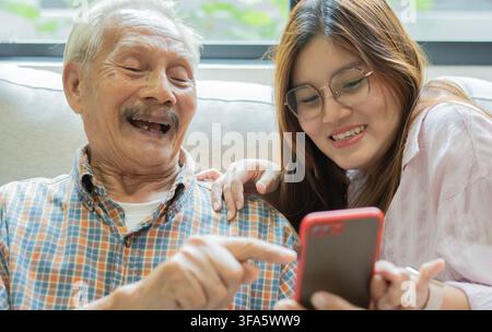 Grand-père et petite-fille heureux assis sur un canapé, se détendant et profitant tout en utilisant un smartphone à la maison. Concept de journée en famille. Banque D'Images