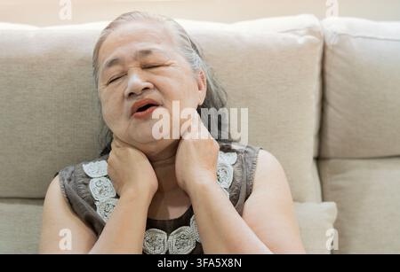 Femme malheureuse de 70 ans reposant sur un canapé tout en massant son cou, souffrant de fortes douleurs au cou à la maison. Concept de problème de santé. Banque D'Images