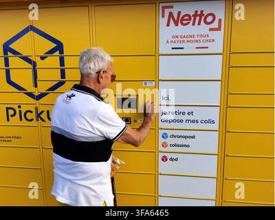 Un homme aux cheveux blancs et aux lunettes se tient devant un casier à colis jaune, utilisant l'écran numérique. Banque D'Images