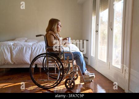Sénior en fauteuil roulant regardant par la fenêtre, soins à domicile et vie indépendante pour les personnes âgées. Récupération et bien-être à domicile, pensif p Banque D'Images