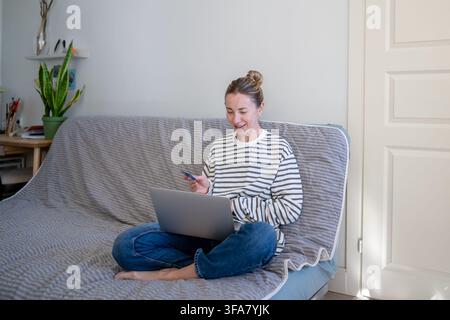 Femme joyeuse achetant en ligne sur un site de commerce électronique depuis chez elle avec un ordinateur portable et une carte de crédit, Banque D'Images