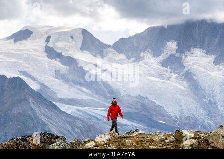 Un randonneur en veste rouge traverse un terrain alpin rocheux avec des glaciers et des pics escarpés derrière lui. La scène se déroule dans les Alpes pennines, Valais, Swi Banque D'Images