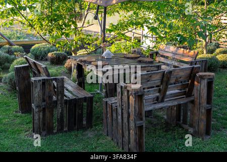 Espace de loisirs en plein air avec table en bois et bancs fabriqués à partir de palettes recyclées, accompagnés de dessous de verre en bois rond posés sur une pelouse verte entourée d'arbres Banque D'Images