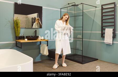 Jeune femme souriante séchant ses cheveux mouillés avec une serviette dans une salle de bain moderne, profitant de sa routine d'hygiène matinale Banque D'Images