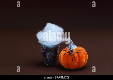 Une tasse sombre en verre en forme de crâne avec un nuage de fumée en coton blanc et duveteux, positionnée à côté d'un citrouille en velours orange fantaisiste équipée de pattes d'araignée noires Banque D'Images