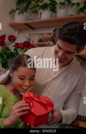 Portrait vertical d'un jeune couple souriant regardant une boîte cadeau en forme de cœur avec des roses rouges en arrière-plan. Banque D'Images