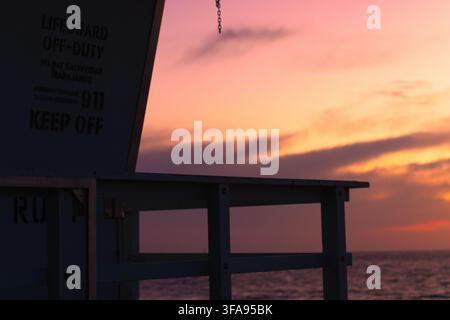 La tour de sauvetage se tient silencieuse au coucher du soleil. Les teintes chaudes du ciel brillent derrière la structure en bois. Le panneau indique «Hors service» et «Interdit d'entrée». L'océan calme reflète une lumière qui s'estompe Banque D'Images
