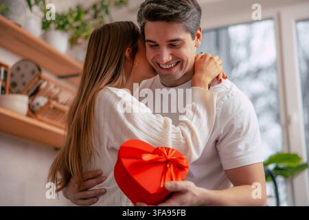 Une femme affectueuse murmure à l'oreille de son partenaire souriant tandis qu'il tient une boîte cadeau rouge en forme de cœur dans une cuisine domestique lumineuse. Banque D'Images