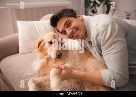 Jeune homme heureux câline un Golden Retriever calme et tient sa patte tout en se relaxant ensemble sur un canapé à la maison. Banque D'Images