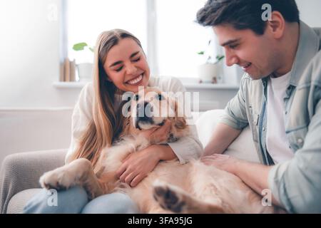 Une femme joyeuse embrasse un Golden Retriever détendu tandis que son partenaire souriant caresse doucement le chien sur un canapé lumineux du salon. Banque D'Images