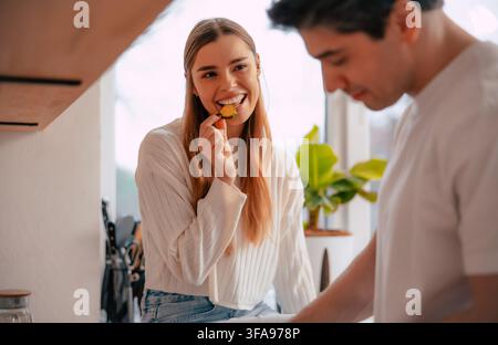 Jeune femme souriante goûte une tranche de courgette fraîche tandis que son partenaire prépare des légumes dans une cuisine moderne et lumineuse Banque D'Images