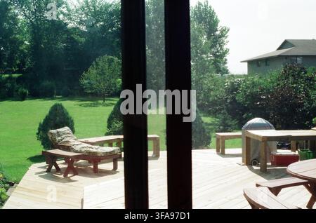 Vue à travers une porte vitrée d'une grande terrasse en bois du jardin arrière avec une chaise longue rembourrée, une station de barbecue et une pelouse verdoyante entourée d'arbres Banque D'Images