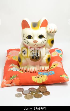 Copilot a dit :   Figurine de chat porte-bonheur Maneki Neko avec pièces japonaises sur un coussin rouge isolé sur fond blanc, Japon Banque D'Images