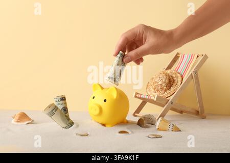 Concept d'épargne de vacances et de fonds de voyage, main mettant un billet de dollar dans une tirelire sur le sable avec des accessoires de plage Banque D'Images