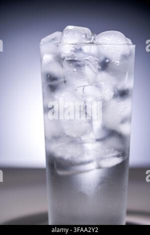 Devant un fond dégradé gris, un grand verre est rempli de glaçons et d'eau glacée, et les parois du verre sont couvertes de gouttes d'eau condensées Banque D'Images