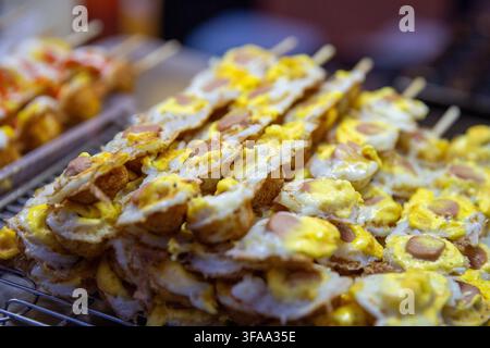 Une vue macro de plusieurs brochettes de street food empilées contenant des œufs de caille frits croustillants garnis de morceaux de saucisse tranchés reposant sur un support métallique chez Jo Banque D'Images