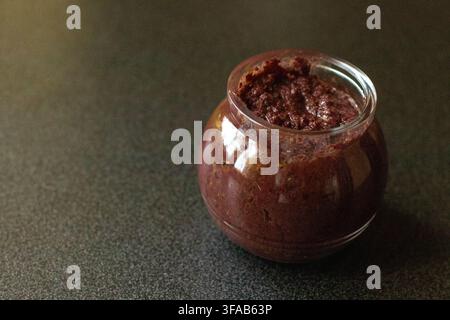 Tapenade d'olives noires dans un pot en verre avec une lumière chaude et dramatique sur une surface sombre tachetée, vue inclinée avec espace de copie à gauche Banque D'Images