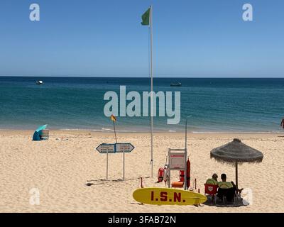 SALEMA, PORTUGAL - 10 juillet 2025 : des maîtres-nageurs veillent depuis une station de plage marquée « I.S.N. » sous un drapeau de sécurité vert sur la plage de sable de Salema Banque D'Images
