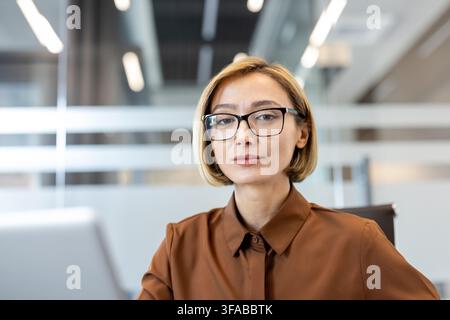 Femme d'affaires professionnelle portant des lunettes assise dans un bureau d'entreprise, regardant directement le spectateur, représentant le leadership, l'expertise et une approche concentrée de sa carrière Banque D'Images