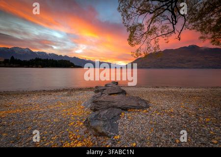 Nouvelle-Zélande - 23 avril 2022 : Vue sur le lac Wakatipu au coucher du soleil avec des nuages orange vifs, une rive rocheuse parsemée de feuilles d'automne et le massif des Remarkables Banque D'Images