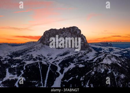 Vue aérienne du Col de Gardena entouré de sommets enneigés et de forêts de pins sous un ciel coucher de soleil vif à Corvara, Trentin-Haut-Adige, Italie. Banque D'Images