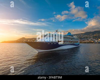 Vue aérienne d'un navire de croisière naviguant dans le port de Funchal au coucher du soleil avec la ville côtière et les montagnes en arrière-plan, Funchal, Madère, Portugal Banque D'Images