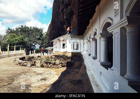 Homme passant devant la façade colonnada blanche du temple des grottes historique de Dambulla, construit sous un immense surplomb rocheux au Sri Lanka Banque D'Images