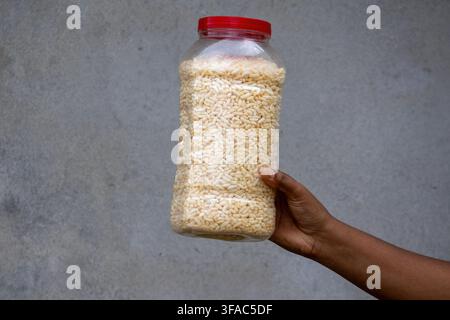 Personne tenant un récipient en plastique rempli de riz soufflé croustillant devant un fond mural gris rustique. Muri traditionnel sud-asiatique conservé pour le thé quotidien Banque D'Images