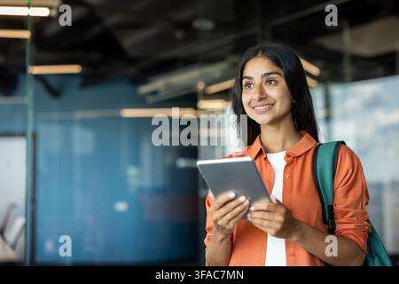 Photo en gros plan d'une jeune étudiante indienne debout à l'intérieur avec un sac à dos, tenant une tablette dans sa main et regardant droit devant. Banque D'Images