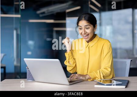 Femme d'affaires indienne heureuse assise au bureau à son lieu de travail, regardant l'écran de son ordinateur portable et se réjouissant de son succès, gesticulant de la main. Banque D'Images