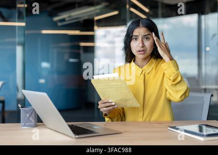 Portrait d'une jeune Indienne inquiète assise à un bureau dans le bureau, tenant une lettre dans une enveloppe et tenant sa tête sous le choc. Banque D'Images